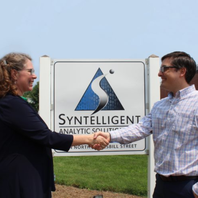 Two people shaking hands in front of Syntelligent sign