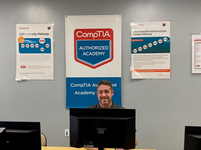 Aaron Winfree sitting behind computer with CompTIA posters in background