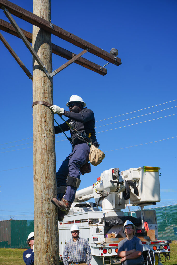 Worker in safety gear climbing a utility pole with a harness while others watch near a bucket truck.