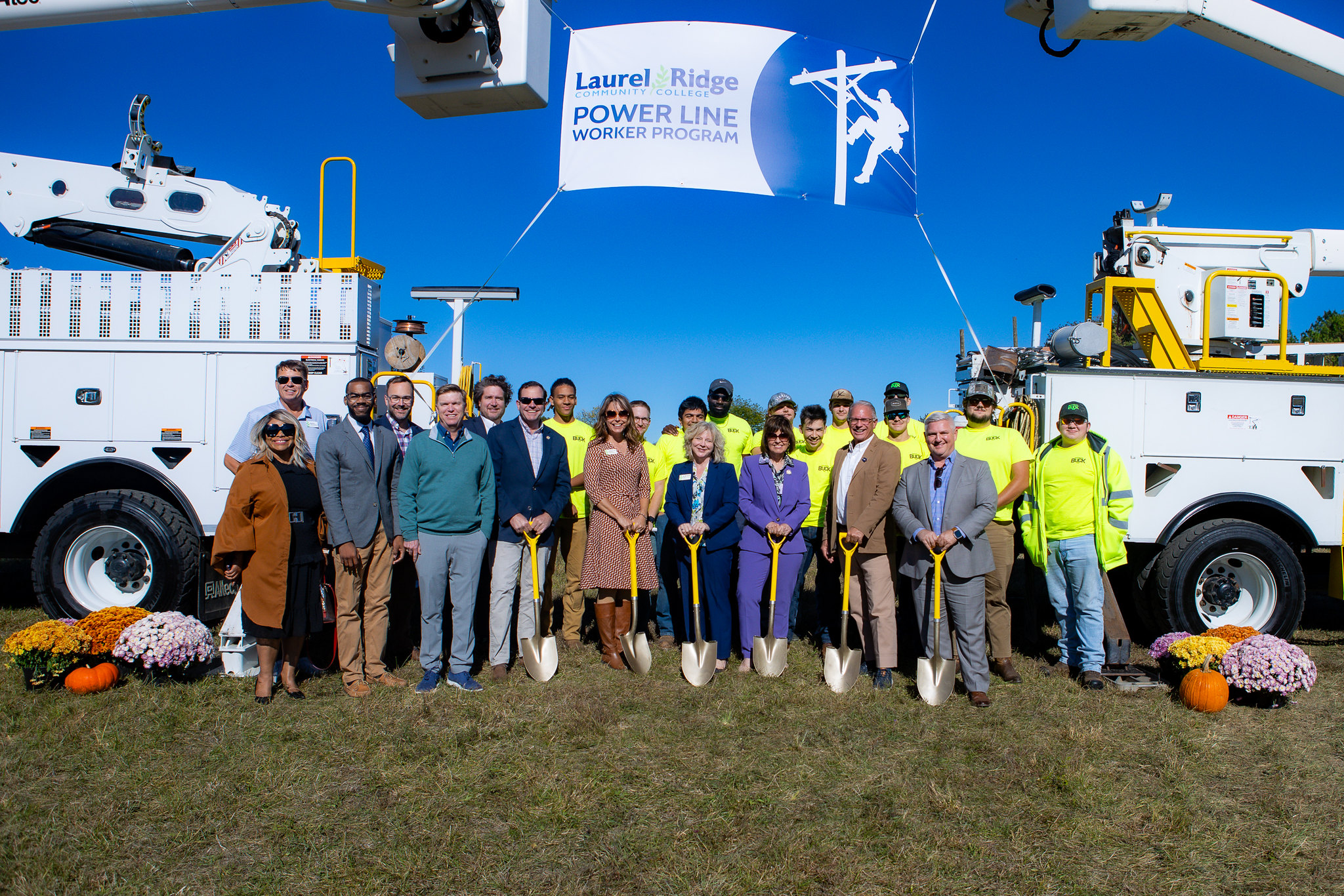 Group of people posing with shovels at a groundbreaking ceremony for the Laurel Ridge Community College Power Line Worker Program, standing between utility trucks under a banner on a clear sunny day.