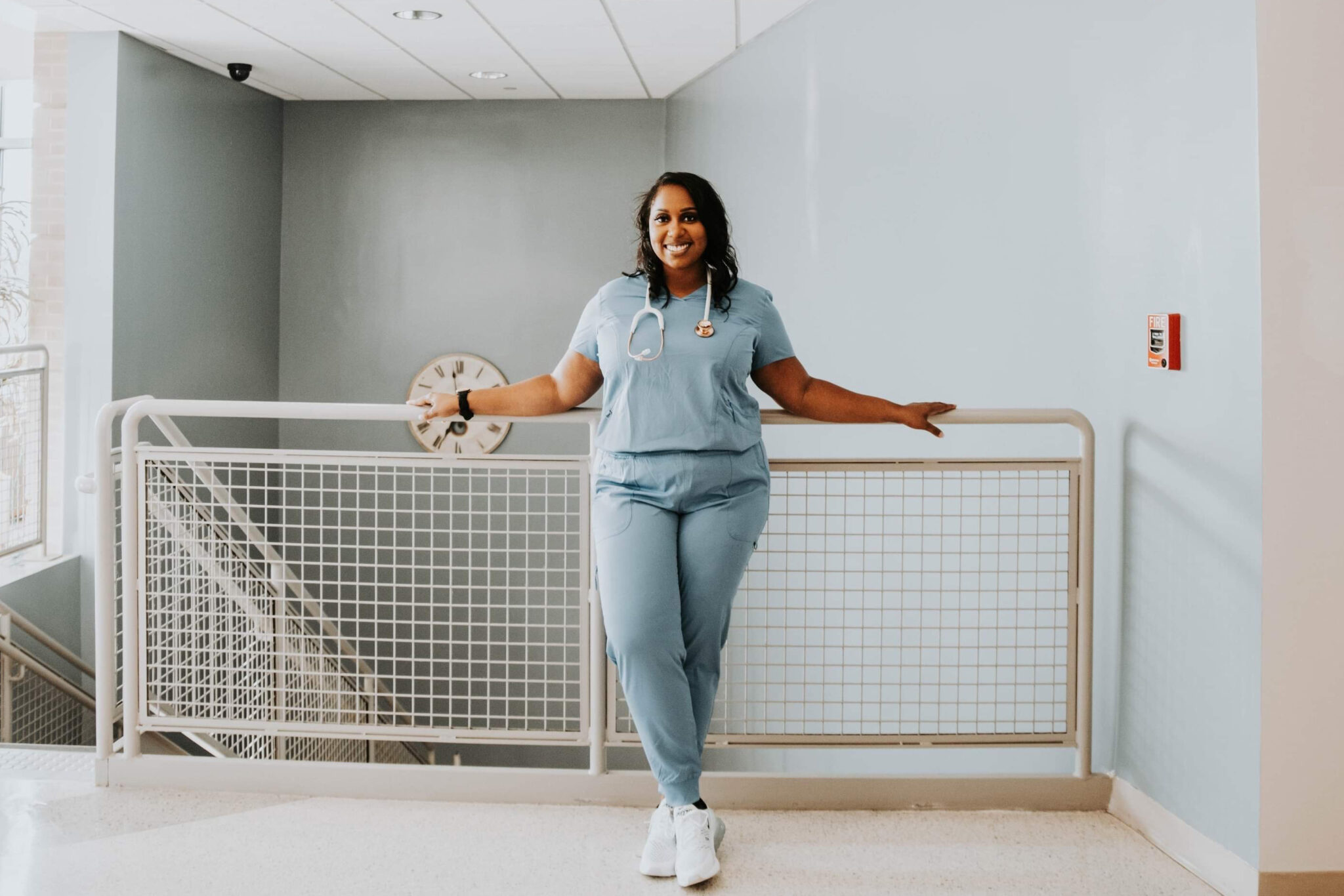 A woman in light blue medical scrubs stands smiling with arms outstretched along a stairway railing, wearing a stethoscope around her neck in a bright, modern indoor setting.