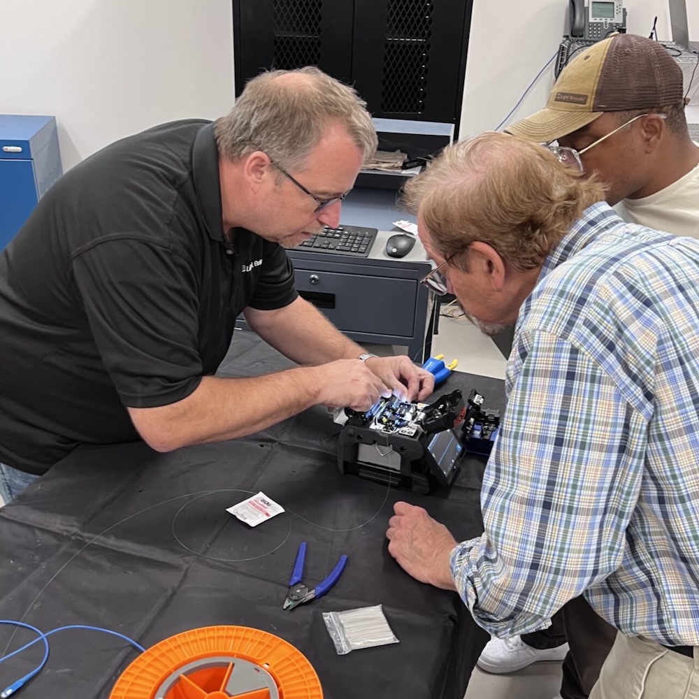 A professor shows two students how to perform installation of a fiber optic cable in the classroom