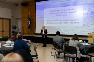 A woman stands on stage presenting at a Laurel Ridge Community College event, with a projected slide behind her that reads, “In the beginning as a Leader: All Work with minimal personal connections. My successful Leadership Journey: Build Connections – Better Together.”