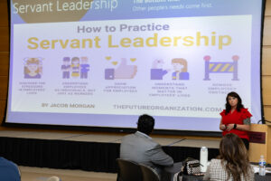 A woman in a red blouse presents at a Laurel Ridge Community College event in front of a slide titled “How to Practice Servant Leadership,” which highlights key principles such as understanding employees, showing appreciation, and removing obstacles.