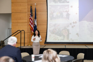 A woman stands in front of a room smiling while talking to a large group