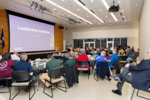 A crowd of people watch a presentation entitled Leadership Institute