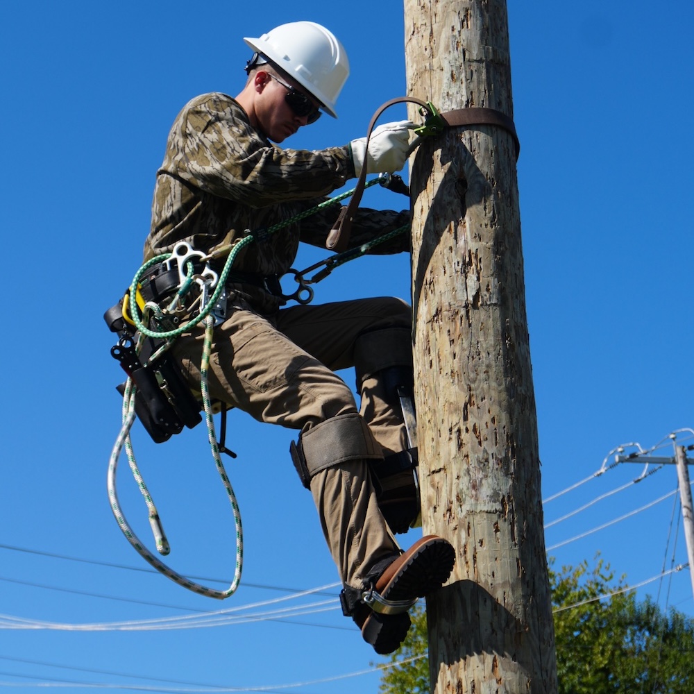 A power line worker climbs up a power line poll while wearing a white hard hat