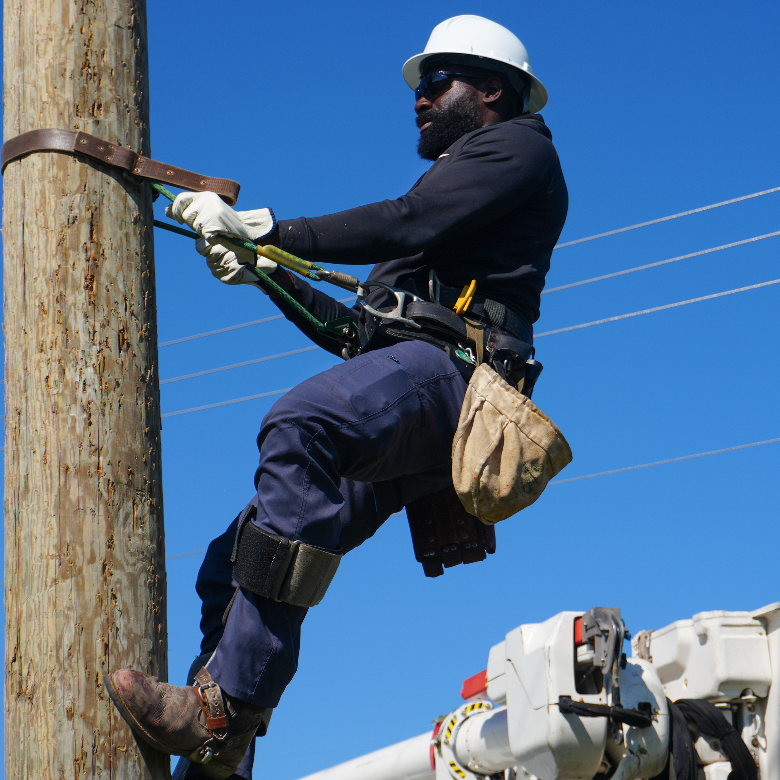 Derrick DeHaney, wearing a white hard hat, safety glasses, gloves, and a tool belt, climbs a wooden utility pole against a clear blue sky while working on electrical lines.