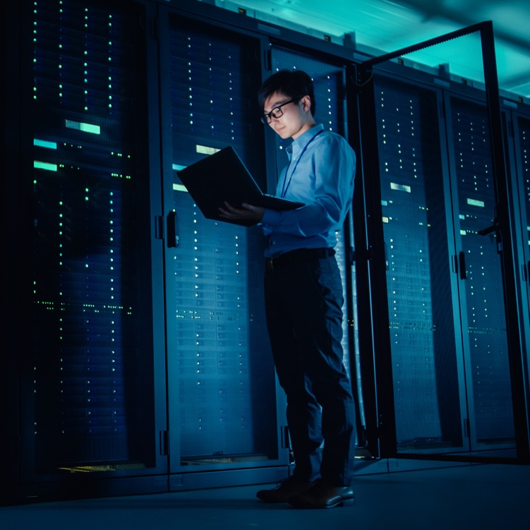 Network and Computer Systems Admin a man looking at a laptop while standing in a dark computer server room
