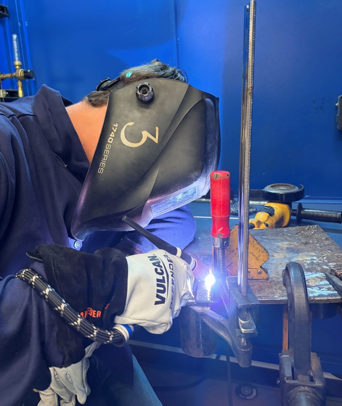 Matthew Del Vacchio welds a metal workpiece while wearing a black 1740 Series welding helmet lowered over his face and a navy flame-resistant jacket. He grips a TIG welding torch with white Vulcan Defender leather gloves, producing a bright electric arc and blue-purple glow. The workpiece is clamped in a welding fixture with a red-handled magnet on a metal workbench, with a yellow DeWalt angle grinder and other tools in the background against a blue booth wall.