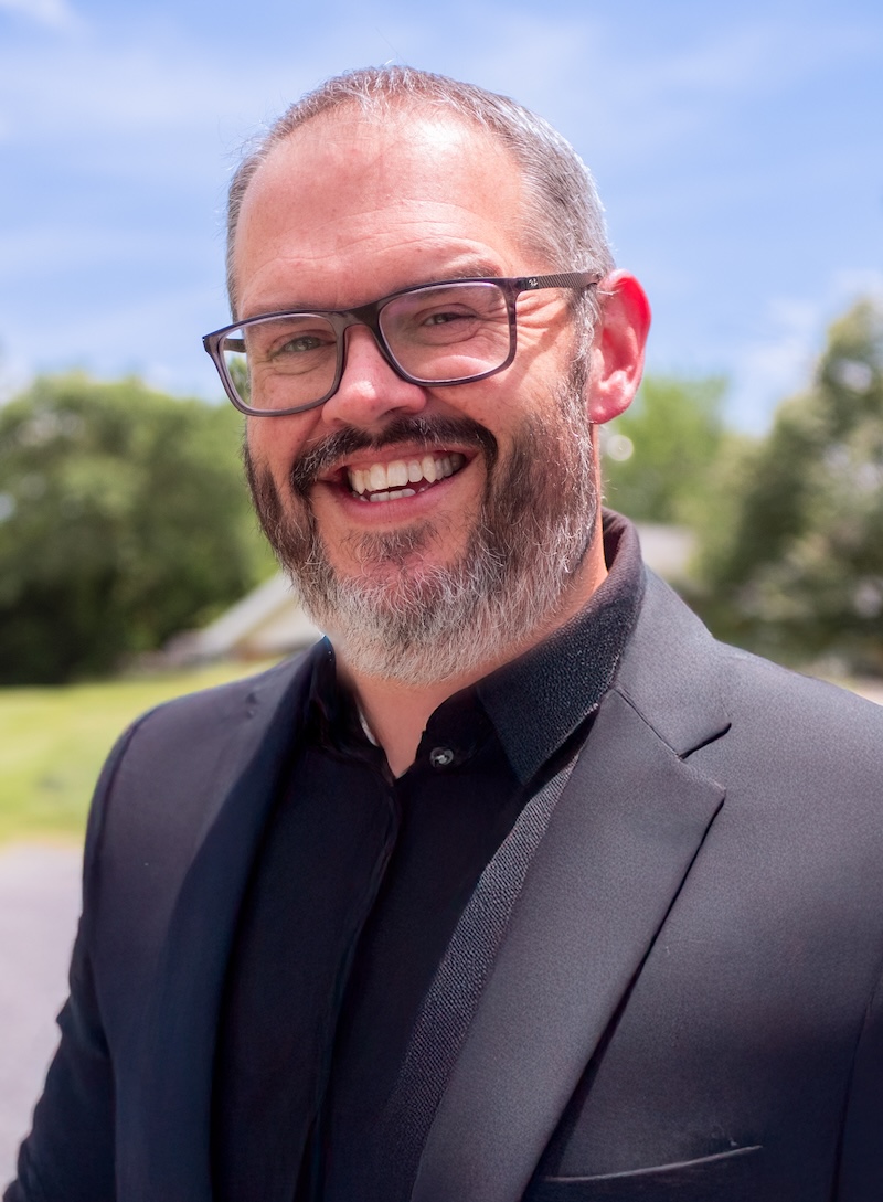 a man in a black blazer and grey / black beard smiling