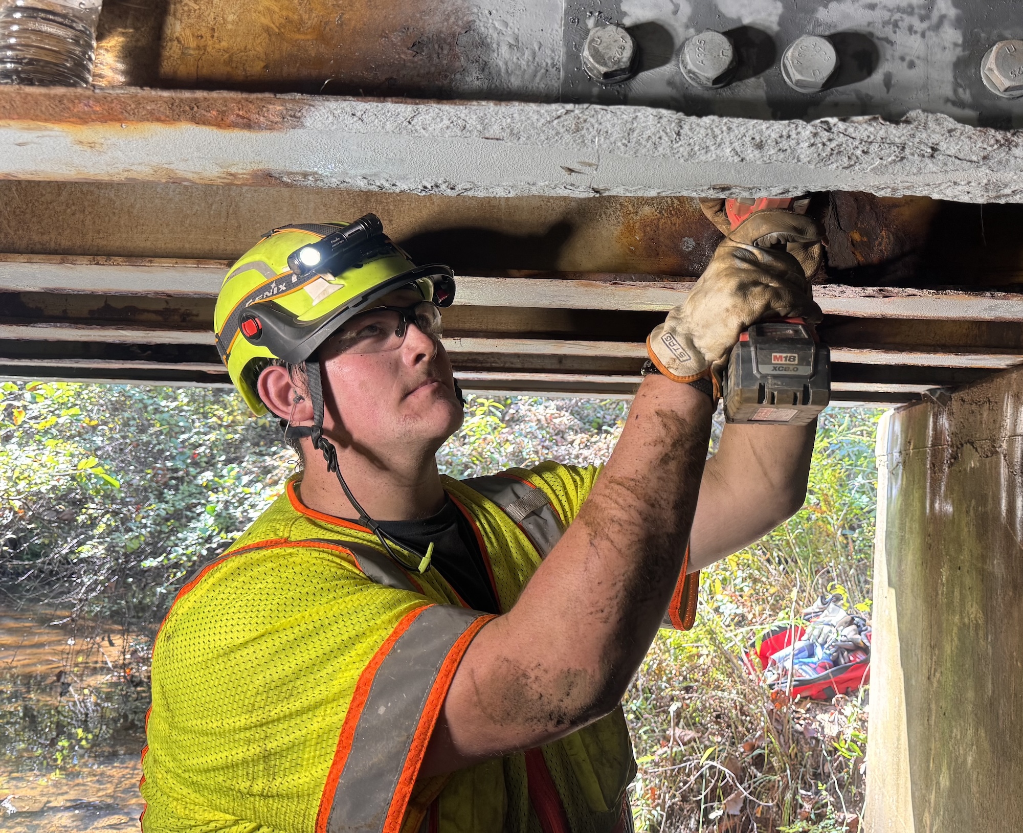 Hunter Jenkins, wearing a hard hat, safety glasses and a high-visibility vest, uses a power drill on the steel underside of a bridge.