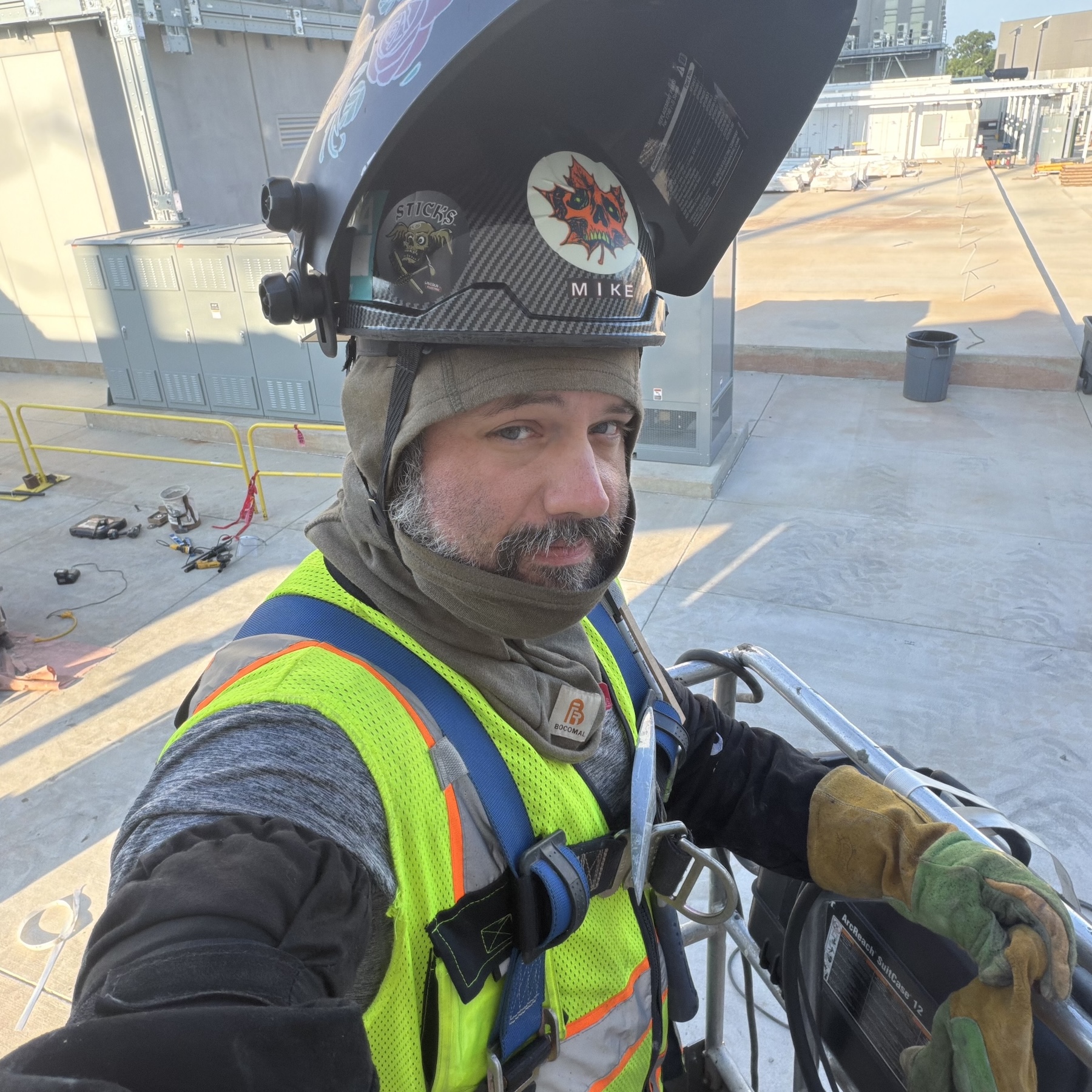 Worker in a safety harness and high-visibility vest takes a selfie from an elevated work platform at an industrial job site while wearing a welding helmet, protective gloves, and cold-weather gear.